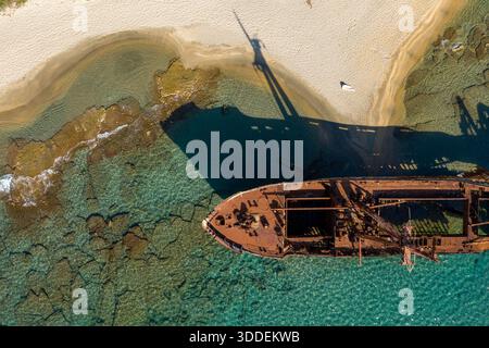 Blick von oben auf das Schiffswrack von Dimitrios, das einen dramatischen Schatten auf goldenen Sand und flaches türkisfarbenes Wasser in der Nähe von Gythio, Griechenland, wirft. Das Sonnenlicht zeigt rostige Metallstrukturen, felsigen Meeresboden und den lebhaften Kontrast zwischen Schiff, Meer und Strand. Stockfoto