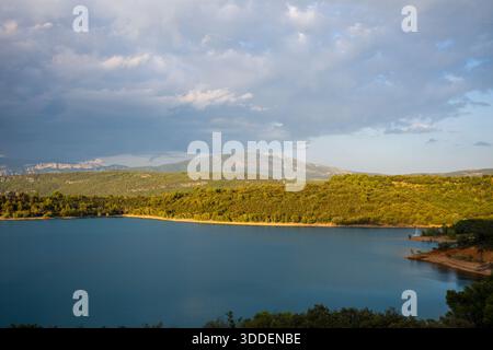 Warmes goldenes Sonnenlicht beleuchtet die sanften grünen Hügel, die das ruhige blaue Wasser des Lac de Sainte-Croix umgeben, mit dramatischen Wolken und fernen Bergen im Hintergrund. Stockfoto