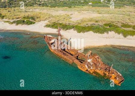 Schräg aus der Vogelperspektive auf das verrostete Dimitrios-Schiffswrack, das in flachen türkisfarbenen Gewässern entlang einer Sandküste in der Nähe von Gythio, Griechenland, gestrandet ist. Das verlassene Schiff steht im Kontrast zum lebhaften Meer und dem natürlichen Grün unter hellem Sonnenlicht. Stockfoto