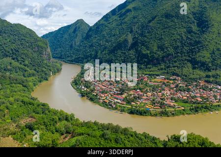 Ein fantastischer Blick aus der Luft auf Nong Khiaw, Laos, zeigt den Fluss Nam ou, der durch ein lebhaftes Dorf schwingt, umgeben von steilen grünen Kalksteinbergen und dichtem tropischen Wald. Die Szene zeigt traditionelle Dächer, üppige Vegetation und die dramatische Landschaft von Nordlaos unter hellem Himmel. Stockfoto