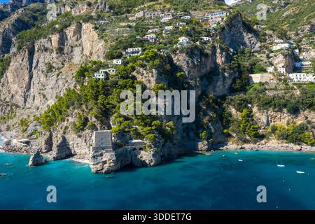 Der historische Wachturm aus Stein steht auf zerklüfteten Klippen über türkisfarbenem Wasser in der Nähe von Positano, Italien. Verstreute Villen am Hügel und üppige mediterrane Vegetation umrahmen die dramatische Küstenlandschaft unter hellem Sonnenlicht. Stockfoto