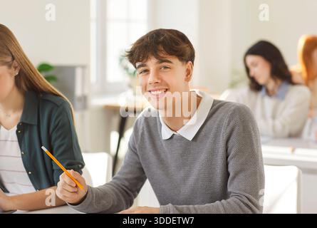 Porträt eines lächelnden jungen Universitätsstudenten, der am Schreibtisch sitzt und Klassenkameraden im Hintergrund studiert. Stockfoto