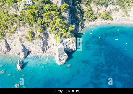 Der historische Wachturm aus Stein steht auf zerklüfteten Klippen über dem türkisfarbenen Mittelmeer in der Nähe von Positano, Italien. Üppige Kiefern und eine felsige Küste umrahmen die dramatische Küste unter hellem Sonnenlicht. Stockfoto