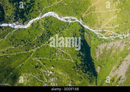 Hochwinkel-Drohnenaufnahme eines schmalen Wildwasserflusses, der sich über üppige grüne Hügel und felsiges Gelände in der Pyrenäenlandschaft kurvt, gespickt mit Wanderwegen und verstreuten Felsbrocken. Stockfoto