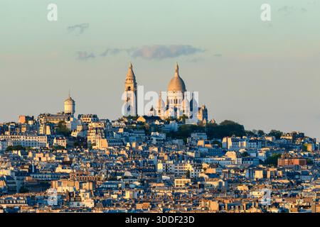 Die ikonische Basilika Sacre Coeur erhebt sich über den dicht gepackten Dächern von Montmartre, die in warmes goldenes Licht bei Sonnenuntergang mit einem weichen, pastellfarbenen Himmel über dem Himmel getaucht sind. Stockfoto