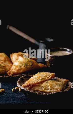 Hausgemachtes türkisches traditionelles Dessert Baklava mit Pistazien serviert auf Keramikplatte mit Nüssen, Tasse Kaffee, Vintage-Cezva auf schwarzem Holztisch. Kopierbereich Stockfoto