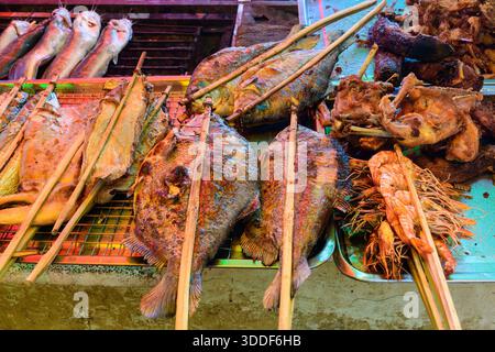 Verschiedene gegrillte Fische, Garnelen und Geflügel werden auf Metalltabletts an einem lebhaften Marktstand in Kambodscha ausgestellt. Das Bild hebt eine Vielzahl von Texturen und Farben unter heller Marktbeleuchtung hervor und zeigt die lokale Street Food-Kultur. Stockfoto