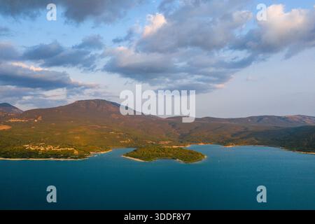 Die warme Abendsonne hebt eine grüne Halbinsel hervor, die in das leuchtend blaue Wasser des Lac de Sainte-Croix ragt, mit sanften Bergen und verstreuten Wolken im Hintergrund. Stockfoto