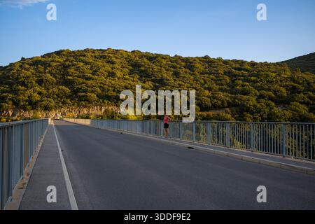 Eine ruhige Straßenbrücke erstreckt sich über ein Tal, das von blauen Geländern begrenzt wird und auf einen üppigen, sonnendurchfluteten Hügel am Lac de Sainte-Croix blickt. Stockfoto