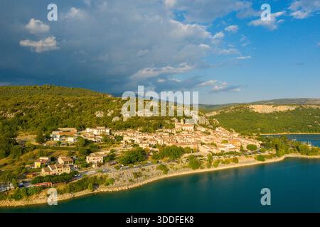 Aus der Vogelperspektive von Bauduen thront über dem lebhaften blauen Wasser des Lac de Sainte-Croix, mit goldenem Sonnenlicht, das durch dramatische Wolken und üppige grüne Hügel im Hintergrund bricht. Stockfoto