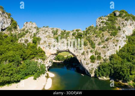 Ein weitläufiger natürlicher Kalksteinbogen erhebt sich über einem sanft geschwungenen Fluss, umgeben von dichtem grünen Wald und zerklüfteten Klippen unter einem klaren blauen Himmel in der Region Ardeche. Stockfoto
