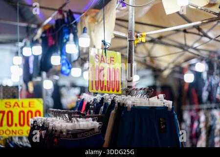 Auf einem belebten Nachtmarkt werden verschiedene Shorts und Kleidung unter hellen Lichtern ausgestellt, mit auffälligen gelben Verkaufsschildern, die die Preise in Landeswährung anzeigen. Die Szene bietet eine lebhafte Atmosphäre im Einzelhandel, beleuchtete Glühbirnen und farbenfrohe Kleidungsstücke in einem Verkaufsstand unter freiem Himmel. Stockfoto
