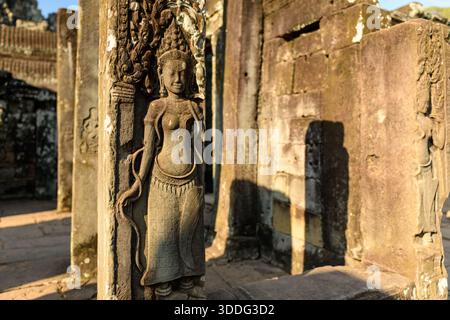 Warmes Sonnenlicht hebt eine aufwändig geschnitzte apsara-Figur auf einer Sandsteinsäule am Bayon-Tempel in Angkor, Kambodscha hervor. Das detaillierte Khmer-Basrelief hebt sich von verwitterten Steinmauern ab und erweckt ein Gefühl alter Kunstfertigkeit und eine ruhige Atmosphäre. Stockfoto