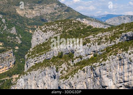 Weitläufiger Blick auf geschichtete Kalksteinklippen, bedeckt mit verstreuten grünen Sträuchern und Bäumen in der Verdon-Schlucht, bei hellem Tageslicht mit weit entfernten Bergkämmen im Hintergrund. Stockfoto