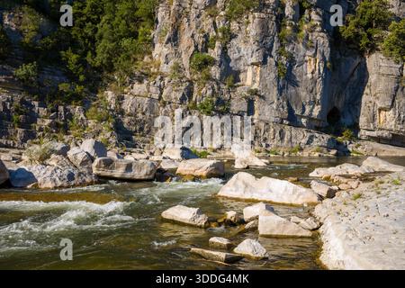 Schnell fließendes Wasser fließt über verstreute weiße Felsen am Fuße einer steilen Kalksteinklippe, mit grünen Flecken und rauem Sonnenlicht in den Gorges de l'Ardeche. Stockfoto