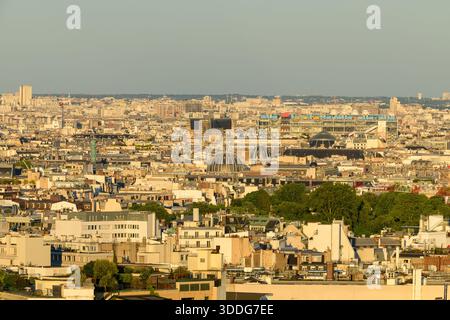 Herrlicher Blick über Paris mit einer dichten Auswahl an Dächern, historischen Kuppeln und dem farbenfrohen Centre Pompidou, alle in sanftes goldenes Abendlicht getaucht. Die städtische Landschaft ist von Grünflächen und geschichteten Architekturformen durchsetzt. Stockfoto