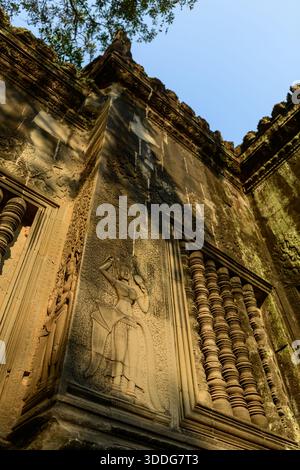 Detaillierter Blick nach oben auf eine Sandsteinmauer in Angkor VAT, Kambodscha, mit einem komplizierten Basreliefs der apsara-Tänzerin und einem traditionellen Balusterfenster. Warmes Sonnenlicht unterstreicht die historischen Khmer-Schnitzereien und die strukturierte Steinoberfläche unter einem klaren Himmel. Stockfoto