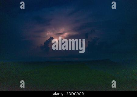 Eine dramatische Gewitterwolke wird von internen Blitzen über schattigen Hügeln unter einem tiefblauen Nachthimmel beleuchtet. Die Szene zeigt mehrschichtige Wolken, subtile Silhouetten und die atmosphärische Spannung eines sich nähernden Sturms. Stockfoto