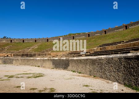 Im römischen Amphitheater in Pompeji, Italien, erhebt sich in geschwungenen Stockwerken alte Stühle. Helles Sonnenlicht hebt das strukturierte vulkanische Mauerwerk und die grasbewachsenen Böschungen unter einem leuchtend blauen Himmel hervor und unterstreicht die historische Größe und das Open-Air-Design des Denkmals. Stockfoto