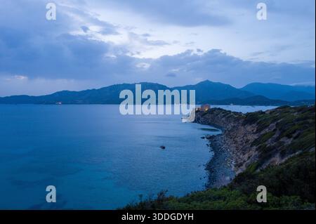 Ruhiger Blick in die Dämmerung auf die zerklüftete Küste von Capo Carbonara mit einer einsamen Festung, die über ruhigem blauem Wasser und fernen Bergsilhouetten unter einem dramatisch bewölkten Himmel beleuchtet wird. Stockfoto