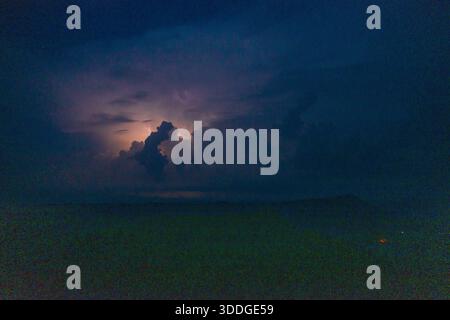 Ein dramatisches Gewitter erleuchtet die Wolken über einem schattigen Bergrücken unter einem tiefblauen Nachthimmel. Die Szene zeigt leuchtende Wolkenformationen, mehrschichtige Silhouetten und eine stimmungsvolle, atmosphärische Landschaft. Stockfoto