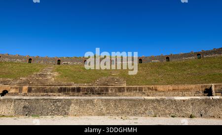 Im römischen Amphitheater in Pompeji, Italien, erhebt sich in geschwungenen Stockwerken alte Stühle. Helles Sonnenlicht hebt das strukturierte vulkanische Mauerwerk und die grasbewachsenen Böschungen unter einem leuchtend blauen Himmel hervor und unterstreicht die historische Größe und das Open-Air-Design des Denkmals. Stockfoto