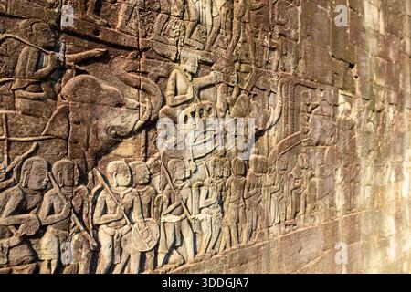Detaillierte Reliefschnitzereien zeigen eine alte Elefantenprozession mit Soldaten und Anhängern an der Sandsteinmauer des Bayon-Tempels in Angkor, Kambodscha. Warmes Sonnenlicht unterstreicht die komplizierte Kunst der Khmer und die strukturierten Steinoberflächen. Stockfoto