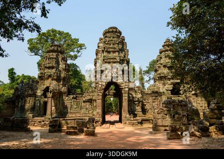 Das Haupttor aus Sandstein und die hohen Türme des Preah Khan Tempels ragen über verstreuten Steinblöcken in Kambodschas Region Angkor. Das Sonnenlicht beleuchtet komplizierte Schnitzereien und verwittertes Mauerwerk, eingerahmt von üppigen tropischen Bäumen und einem klaren Himmel. Stockfoto