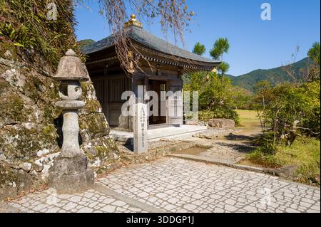 Kleiner Holzschrein mit einer Steinlaterne steht neben moosigen Felsen und Grün unter klarem blauem Himmel in Kumano-Nachi, Japan. Helles Sonnenlicht hebt die Texturen von Holz, Stein und umgebendem Laub hervor. Stockfoto