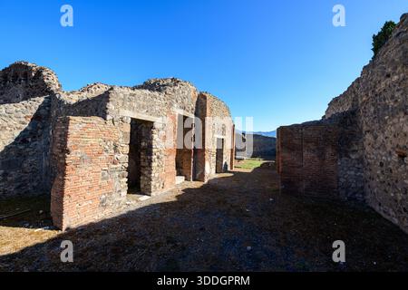 Alte Ziegelmauern und Steinmauern bilden die Überreste des Hauses Cornelli in Pompeji, Italien. Helles Sonnenlicht und tiefe Schatten unterstreichen das strukturierte Mauerwerk unter einem leuchtend blauen mediterranen Himmel. Stockfoto