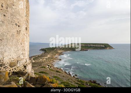 Der historische Wachturm aus Stein erhebt sich über der dramatischen Küste von Capo San Marco, Sardinien, mit gewundenen unbefestigten Wegen und strukturierten Klippen, die unter einem gedämpften Himmel auf das ruhige Mittelmeer treffen. Stockfoto
