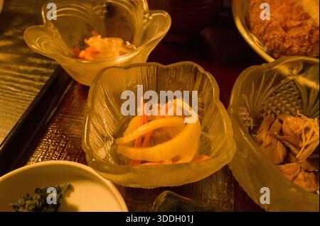 Nahaufnahme traditioneller japanischer Beilagen, die in strukturierten Glasschalen auf einem lackierten Tablett serviert werden, mit eingelegtem Gemüse und gekochten Speisen unter warmer Beleuchtung in Hongu, Japan. Stockfoto