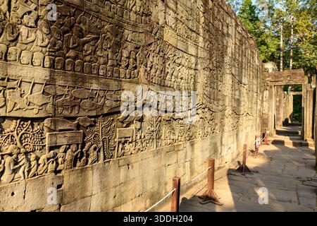 Das warme Nachmittagslicht beleuchtet die komplizierten Flachreliefs an einer Sandsteinwand am Bayon Tempel in Angkor, Kambodscha. Die detailreiche Szene zeigt Figuren, Boote und Tiere mit strukturierten Oberflächen und einem Korridor, der in die antiken Ruinen führt. Stockfoto