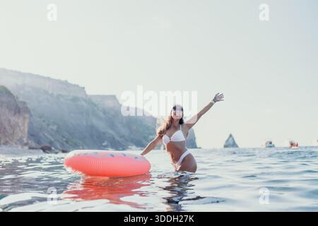 Frau, Meer, Spaß: Fröhliche Frau in weißem Bikini steht im Meer mit rosa Donut Ring und genießt Sommerurlaub. Stockfoto