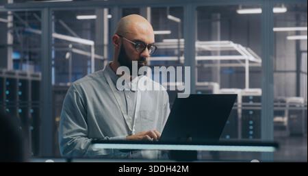 Fokussierter IT-Techniker mit Laptop in der Serverhalle. Mann in leichtem Shirt steht und arbeitet intensiv, Hintergrund zeigt Server-Racks modernes Rechenzentrum. Konzept vor-Ort-Diagnose und Systemwartung. Stockfoto