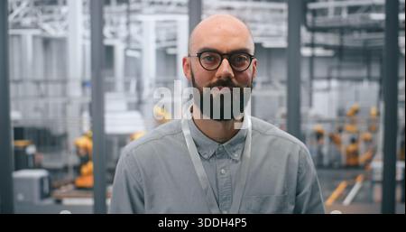 Engagierter männlicher technischer Spezialist posiert selbstbewusst vor einem automatisierten Industriewerk mit Maschinen. Konzept der Rolle bei der Verwaltung von Robotik und fortschrittlichen Fertigungsprozessen. Hochformat. Stockfoto