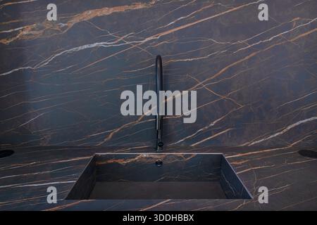 Modernes schwarzes Spülbecken mit passenden matten Armaturen, vor einem dunklen Hintergrund in Marmor-Optik und einer Arbeitsfläche mit dezenter brauner Aderung Stockfoto