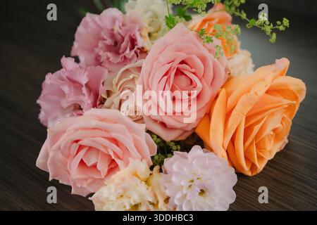 Blumenarrangements mit zarten rosa und pfirsichfarbenen Rosen und frischen grünen Akzenten, elegant auf dunkler Holzoberfläche angeordnet, schöner Blumenstrauß für Hochzeiten Stockfoto