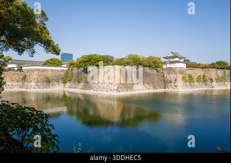 Ein traditioneller japanischer Burgturm erhebt sich über massiven Steinmauern und einem ruhigen Graben, umgeben von üppigem Grün unter einem klaren blauen Himmel in Osaka. Reflexionen tanzen auf dem ruhigen Wasser und schaffen eine friedliche und zeitlose Atmosphäre. Stockfoto