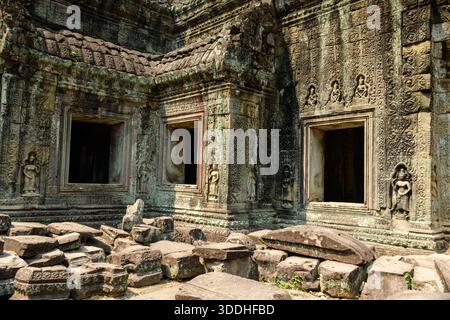 Die verwitterten Sandsteinwände des Preah Khan Tempels in Kambodschas Angkor zeigen komplizierte Schnitzereien, Basreliefs und offene Fenster. Sonnenlicht beleuchtet die detailreiche Fassade und die moosbedeckten Steine, die über den alten Innenhof verstreut sind. Stockfoto