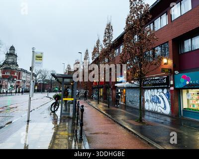 Manchester, Großbritannien - 06. Dezember 2025: Regnerische Manchester Street mit Bushaltestelle, Radfahrern, Graffitis und Geschäften; städtisches Flair. Stockfoto