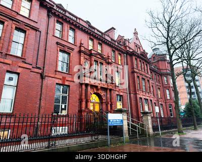 Manchester, Großbritannien - 06. Dezember 2025: Historisches rotes Backsteingebäude in Manchester mit einem bogenförmigen Eingang, einem Eisenzaun und klassischer Architektur. Stockfoto