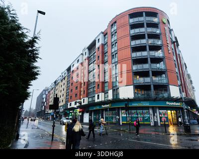 Manchester, Großbritannien - 06. Dezember 2025: Regentag in Manchester zeigt einen modernen Wohnblock mit Geschäften und Fußgängern. Stockfoto