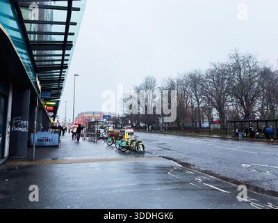 Manchester, Großbritannien - 06. Dezember 2025: Regnerische Manchester Street mit Fahrrädern, Shoppern und Geschäften entlang einer nassen Stadtpromenade. Stockfoto
