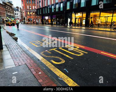 Manchester, Großbritannien - 01. Dezember 2025: Die regnerische Stadtstraße in Manchester zeigt heute eine Bushaltestelle, Radfahrer und Glasfassaden. Stockfoto
