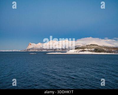 Winter Küstenpanorama Norwegens mit schneebedeckten Bergen, niedrigen Wolken und einem tiefblauen Meer unter stimmungsvollem Licht. Stockfoto