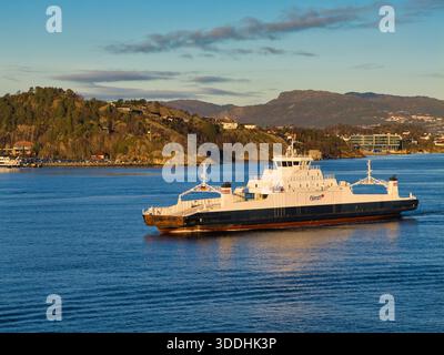 Bergen, Norwegen - 25. November 2025: Eine Autofähre fährt über den blauen Fjord in Bergen, Norwegen, mit Häusern und Bergen in Hügeln und im milden Herbstlicht. Stockfoto