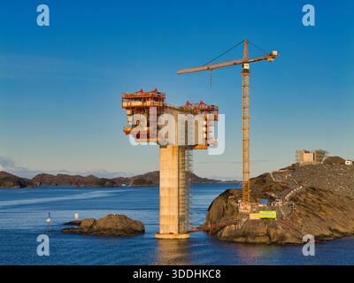 Küste, Norwegen - 25. November 2025: Betonbrücke und Turmkran erheben sich über dem blauen Meer an der norwegischen Küste und fangen den modernen Tiefbau zur goldenen Stunde ein. Stockfoto