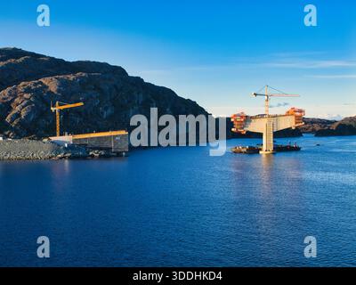 Küste, Norwegen - 25. November 2025: Brückenpfeiler erheben sich aus blauem Wasser, während Kräne an einem Küstenübergang entlang der norwegischen Küste arbeiten, eingerahmt von zerklüfteten Felsen und klarem Himmel. Stockfoto