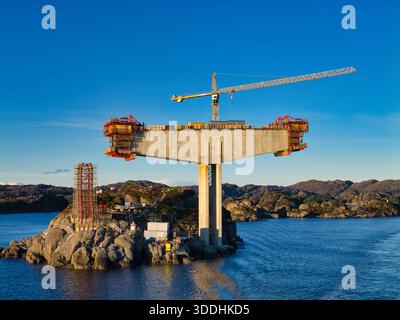 Küste, Norwegen - 25. November 2025: Betonbrücke mit Schalung und Turmkran erhebt sich über felsigen Inselchen an der norwegischen Küste, gefangen in hellem Winterlicht und klarem blauem Himmel. Stockfoto